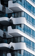 A modern building facade with a series of curved balconies, glass panels, and a sleek architectural design. Trees are placed on the balconies, adding a touch of greenery to the urban structure.