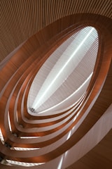An abstract architectural perspective of a structure with curved wooden and metallic elements forming a spiraling, upward trajectory towards a skylight. Light passes through the opening, creating a dynamic play of shadows and highlights along the layers.