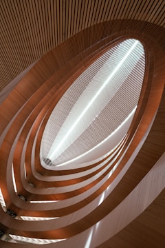 An abstract architectural perspective of a structure with curved wooden and metallic elements forming a spiraling, upward trajectory towards a skylight. Light passes through the opening, creating a dynamic play of shadows and highlights along the layers.