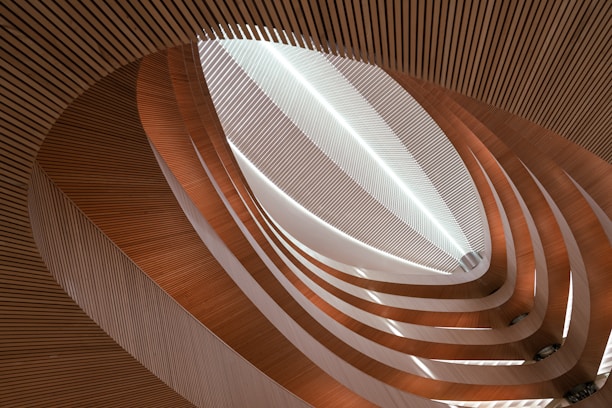 An abstract architectural interior featuring a series of curved, layered wooden slats creating an intricate, flowing pattern. The design is highlighted by soft and natural lighting, creating a sense of depth and dimension.