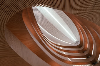 An abstract architectural interior featuring a series of curved, layered wooden slats creating an intricate, flowing pattern. The design is highlighted by soft and natural lighting, creating a sense of depth and dimension.