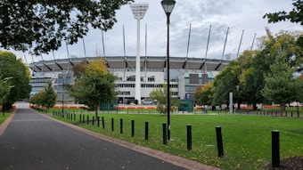 A large sports stadium with a modern design is surrounded by lush green trees and a spacious lawn. The foreground features a pathway lined with evenly spaced black posts, while a tall light pole stands prominently in front of the stadium. The sky is overcast, creating a contrast with the vibrant greenery.