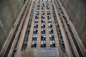 A towering building with numerous windows and air conditioning units. The structure is made of concrete and appears to be part of an urban setting. The words 'Criminal Courts Building' are visible at the base of the building.