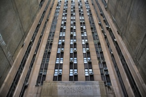 A towering building with numerous windows and air conditioning units. The structure is made of concrete and appears to be part of an urban setting. The words 'Criminal Courts Building' are visible at the base of the building.