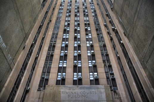 A towering building with numerous windows and air conditioning units. The structure is made of concrete and appears to be part of an urban setting. The words 'Criminal Courts Building' are visible at the base of the building.