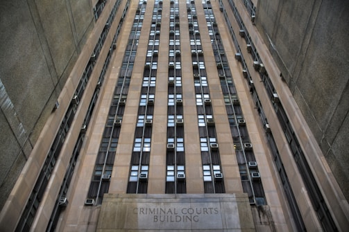 A towering building with numerous windows and air conditioning units. The structure is made of concrete and appears to be part of an urban setting. The words 'Criminal Courts Building' are visible at the base of the building.