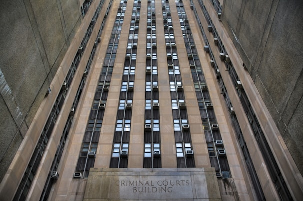 A towering building with numerous windows and air conditioning units. The structure is made of concrete and appears to be part of an urban setting. The words 'Criminal Courts Building' are visible at the base of the building.