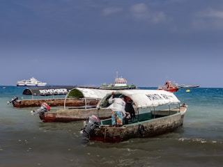 Several small fishing boats with canopies are docked in shallow waters near a beach. One of the boats has the word 'REMEMBER' written on its canopy, while another boat has 'MAKUN TOUR' written on it. A person is standing in one of the boats, partially inside the canopy. In the background, there are a few larger boats and ships on the blue ocean under a clear sky.