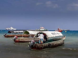 Several small fishing boats with canopies are docked in shallow waters near a beach. One of the boats has the word 'REMEMBER' written on its canopy, while another boat has 'MAKUN TOUR' written on it. A person is standing in one of the boats, partially inside the canopy. In the background, there are a few larger boats and ships on the blue ocean under a clear sky.