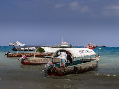 Several small fishing boats with canopies are docked in shallow waters near a beach. One of the boats has the word 'REMEMBER' written on its canopy, while another boat has 'MAKUN TOUR' written on it. A person is standing in one of the boats, partially inside the canopy. In the background, there are a few larger boats and ships on the blue ocean under a clear sky.