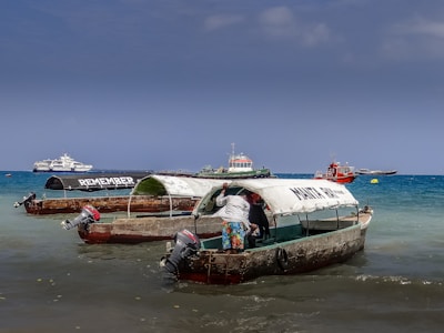 Several small fishing boats with canopies are docked in shallow waters near a beach. One of the boats has the word 'REMEMBER' written on its canopy, while another boat has 'MAKUN TOUR' written on it. A person is standing in one of the boats, partially inside the canopy. In the background, there are a few larger boats and ships on the blue ocean under a clear sky.