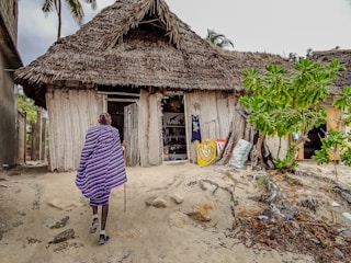 A person in traditional attire walks towards a rustic building with a thatched roof, surrounded by sand and vegetation. The entrance features artwork, including a lion painting, and the walls are constructed with natural materials.