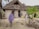 A person in traditional attire walks towards a rustic building with a thatched roof, surrounded by sand and vegetation. The entrance features artwork, including a lion painting, and the walls are constructed with natural materials.