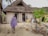 A person in traditional attire walks towards a rustic building with a thatched roof, surrounded by sand and vegetation. The entrance features artwork, including a lion painting, and the walls are constructed with natural materials.