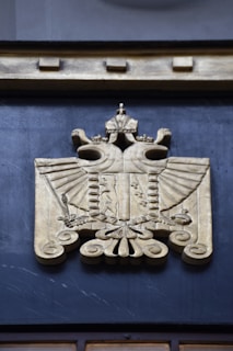 A detailed relief carving of an eagle with outspread wings, adorned with a crown and intricate designs. The eagle is symmetrical, and the carving is done in a golden hue. The artistic style is ornate, featuring numerous decorative elements.