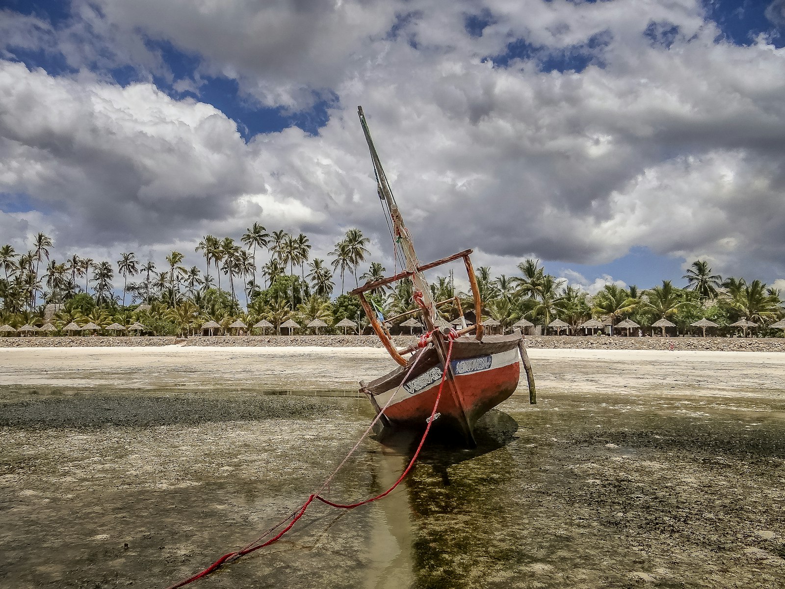 A wooden dhow resting on the exposed seabed at low tide, with palms and thatched shades beyond