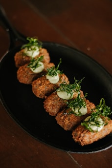 A black skillet contains six golden-brown croquettes arranged in a row. Each croquette is topped with a dollop of creamy green sauce and garnished with finely chopped green herbs.