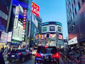 A bustling urban street scene featuring numerous bright, colorful advertisements and digital billboards on high-rise buildings. Several cars are on the street, and pedestrians are visible on the sidewalks. The scene suggests a vibrant city life.