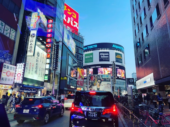 A bustling urban street scene featuring numerous bright, colorful advertisements and digital billboards on high-rise buildings. Several cars are on the street, and pedestrians are visible on the sidewalks. The scene suggests a vibrant city life.