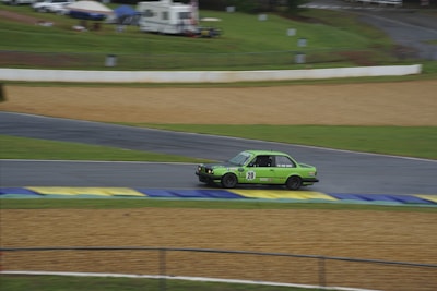 A green racing car with the number 20 is moving quickly along a race track. The track is surrounded by grassy areas and patches of gravel. Tents and mobile homes can be seen in the background, indicating a racing event or festival atmosphere.
