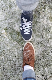 Two shoes touching toe-to-toe on a snow-dusted grassy surface. One shoe is black with white stripes, and the other is brown with visible snow on top.