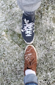 Two shoes touching toe-to-toe on a snow-dusted grassy surface. One shoe is black with white stripes, and the other is brown with visible snow on top.