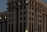 A large, multi-story building with a modern architectural design. The facade is structured with repeated rectangular windows and surfaces, illuminated by external lights that create a warm glow against the darkening sky. The building's exterior is clad in light-colored stone or concrete panels.