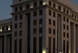 A large, multi-story building with a modern architectural design. The facade is structured with repeated rectangular windows and surfaces, illuminated by external lights that create a warm glow against the darkening sky. The building's exterior is clad in light-colored stone or concrete panels.