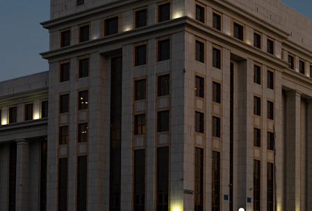 A large, multi-story building with a modern architectural design. The facade is structured with repeated rectangular windows and surfaces, illuminated by external lights that create a warm glow against the darkening sky. The building's exterior is clad in light-colored stone or concrete panels.