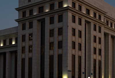 A large, multi-story building with a modern architectural design. The facade is structured with repeated rectangular windows and surfaces, illuminated by external lights that create a warm glow against the darkening sky. The building's exterior is clad in light-colored stone or concrete panels.