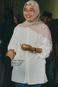 A person wearing a beige headscarf and glasses, dressed in a white shirt, is smiling warmly while interacting with someone holding a plate. The background features a few other individuals.
