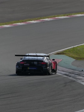 A race car is navigating a track, surrounded by a paved racing surface and bordered by grassy and painted areas. The vehicle is a sports car with visible sponsors' logos and a number on its body, suggesting a competitive racing event.