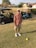 An individual is standing on a golf course, holding a golf club. He is wearing a cap, sunglasses, a short-sleeved shirt, shorts, and golf shoes. Two golf carts are visible in the background, parked on the edge of the grass. The setting includes residential houses and some trees.