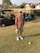 An individual is standing on a golf course, holding a golf club. He is wearing a cap, sunglasses, a short-sleeved shirt, shorts, and golf shoes. Two golf carts are visible in the background, parked on the edge of the grass. The setting includes residential houses and some trees.