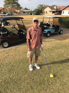 An individual is standing on a golf course, holding a golf club. He is wearing a cap, sunglasses, a short-sleeved shirt, shorts, and golf shoes. Two golf carts are visible in the background, parked on the edge of the grass. The setting includes residential houses and some trees.