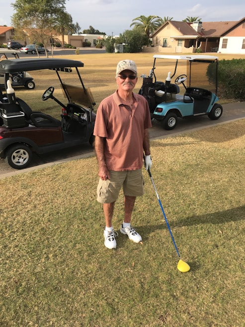 An individual is standing on a golf course, holding a golf club. He is wearing a cap, sunglasses, a short-sleeved shirt, shorts, and golf shoes. Two golf carts are visible in the background, parked on the edge of the grass. The setting includes residential houses and some trees.