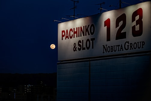 A nighttime scene with a large illuminated sign reading 'Pachinko & Slot 123 Nobuta Group' on the side of a building. A full moon is visible in the dark blue sky, adding a serene touch to the urban environment. The silhouette of buildings can be seen in the background.