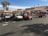 Several golf carts are parked in a parking lot under a clear blue sky. People are gathered around and sitting in some of the golf carts, which appear to be parked in front of a building with a red-tiled roof. There are palm trees in the background, and it looks like a sunny day.