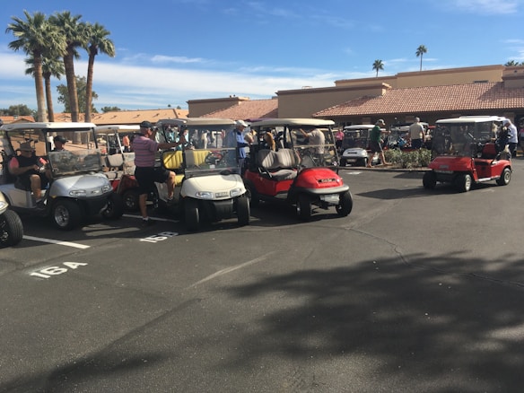 Several golf carts are parked in a parking lot under a clear blue sky. People are gathered around and sitting in some of the golf carts, which appear to be parked in front of a building with a red-tiled roof. There are palm trees in the background, and it looks like a sunny day.