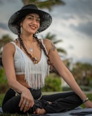 A person with braided hair is sitting cross-legged outdoors. They are wearing a wide-brimmed black hat, a white fringed top, and black pants. Large earrings, necklaces, and tattoos are visible. The background consists of a blurred palm tree and a cloudy sky, suggesting a tropical or beach setting. The person is smiling and appears relaxed.