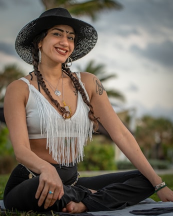 A person with braided hair is sitting cross-legged outdoors. They are wearing a wide-brimmed black hat, a white fringed top, and black pants. Large earrings, necklaces, and tattoos are visible. The background consists of a blurred palm tree and a cloudy sky, suggesting a tropical or beach setting. The person is smiling and appears relaxed.