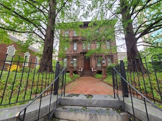 A large, historic stone building with classic architectural features, framed by two large trees. The building is accessed by a brick pathway and steps leading up to a grand wooden door. Wrought iron fencing and lush green foliage surround the front garden area.