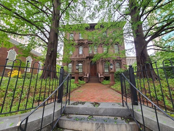 A large, historic stone building with classic architectural features, framed by two large trees. The building is accessed by a brick pathway and steps leading up to a grand wooden door. Wrought iron fencing and lush green foliage surround the front garden area.