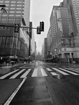 A black and white cityscape photograph of an urban street scene with high-rise buildings lining both sides. The street is wet, suggesting recent rain, and features a visible zebra crossing. Traffic signals and street signs are visible overhead, with a few pedestrians carrying umbrellas.