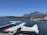 A seaplane with white and orange colors is docked at a wooden pier on a vast body of water. In the background, there are lush green trees lining the opposite shore, with mountains rising beyond them under a clear blue sky. A small boat can be seen on the water, and a red building is situated on the pier to the right of the image.