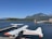 A seaplane with white and orange colors is docked at a wooden pier on a vast body of water. In the background, there are lush green trees lining the opposite shore, with mountains rising beyond them under a clear blue sky. A small boat can be seen on the water, and a red building is situated on the pier to the right of the image.