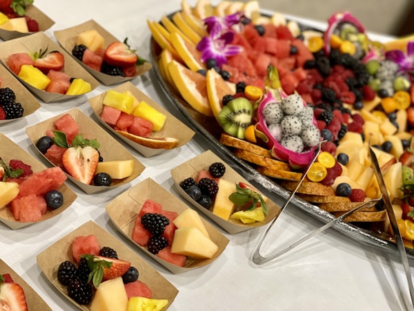 An assortment of fruit pieces arranged elegantly on a large platter and in small paper boats. The spread includes watermelon, pineapple, strawberries, blackberries, raspberries, kiwi, dragon fruit, blueberries, and melon. Decorative orchid flowers and citrus fruits add a colorful touch. The arrangement is festive and inviting.
