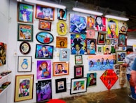 A vibrant collection of framed artworks displayed on a gallery wall. The pieces range from colorful portraits to various artistic styles and themes. Each artwork showcases unique characters or figures, while the frames vary in shape and size. The setting appears lively and creative, with another person standing on the right side.