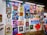 A vibrant collection of framed artworks displayed on a gallery wall. The pieces range from colorful portraits to various artistic styles and themes. Each artwork showcases unique characters or figures, while the frames vary in shape and size. The setting appears lively and creative, with another person standing on the right side.