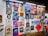 A vibrant collection of framed artworks displayed on a gallery wall. The pieces range from colorful portraits to various artistic styles and themes. Each artwork showcases unique characters or figures, while the frames vary in shape and size. The setting appears lively and creative, with another person standing on the right side.